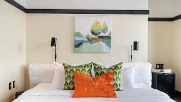 Una cama bien hecha con sábanas blancas y dos almohadas, una estampada en verde y otra en naranja, en una habitación de hotel con un cuadro de un paisaje en la pared y una mesita de noche negra con una taza y un platillo.