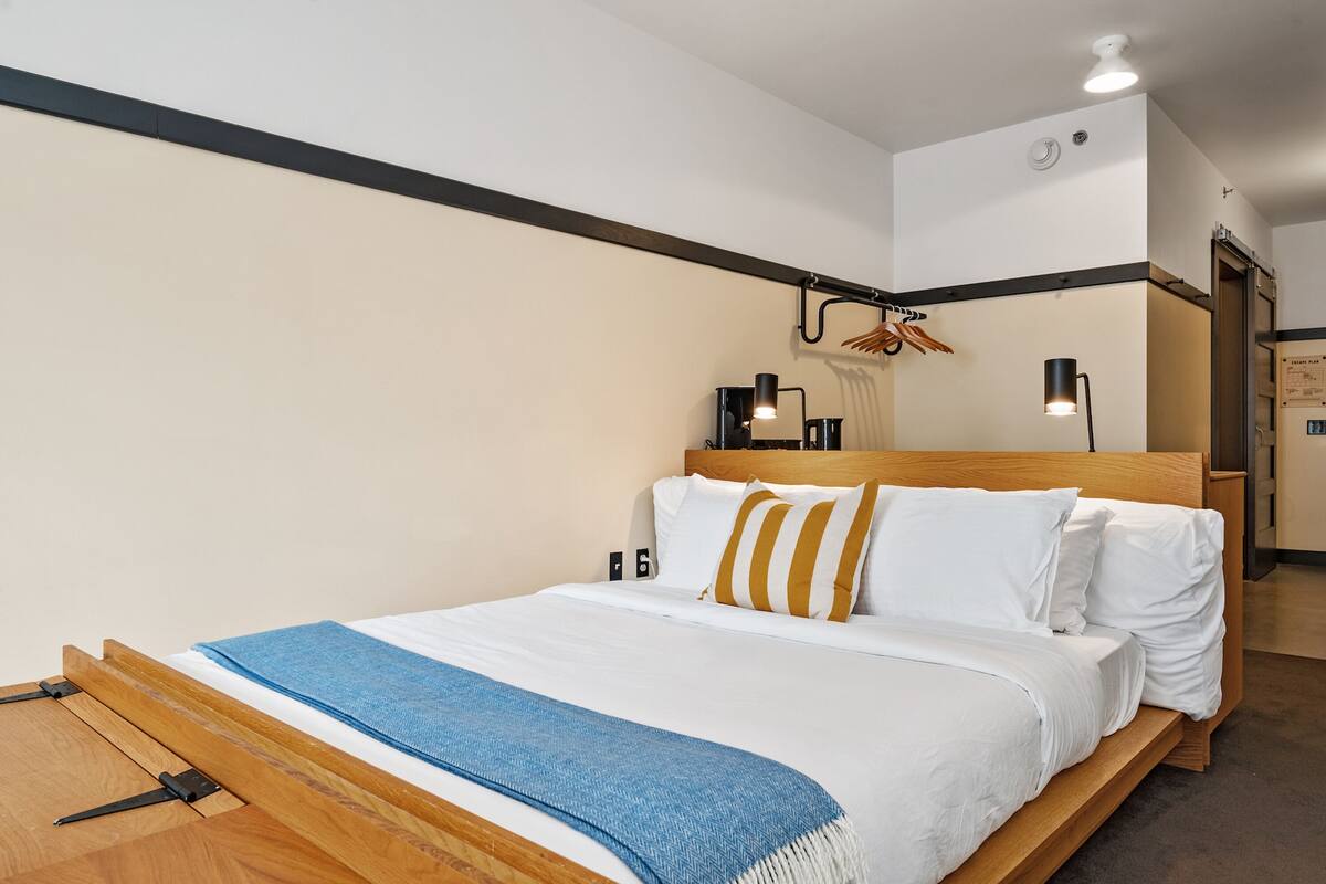 A neatly made bed with a wooden headboard, a blue throw blanket, and white bedding. There are two wall-mounted lamps and a coat rack with clothes.