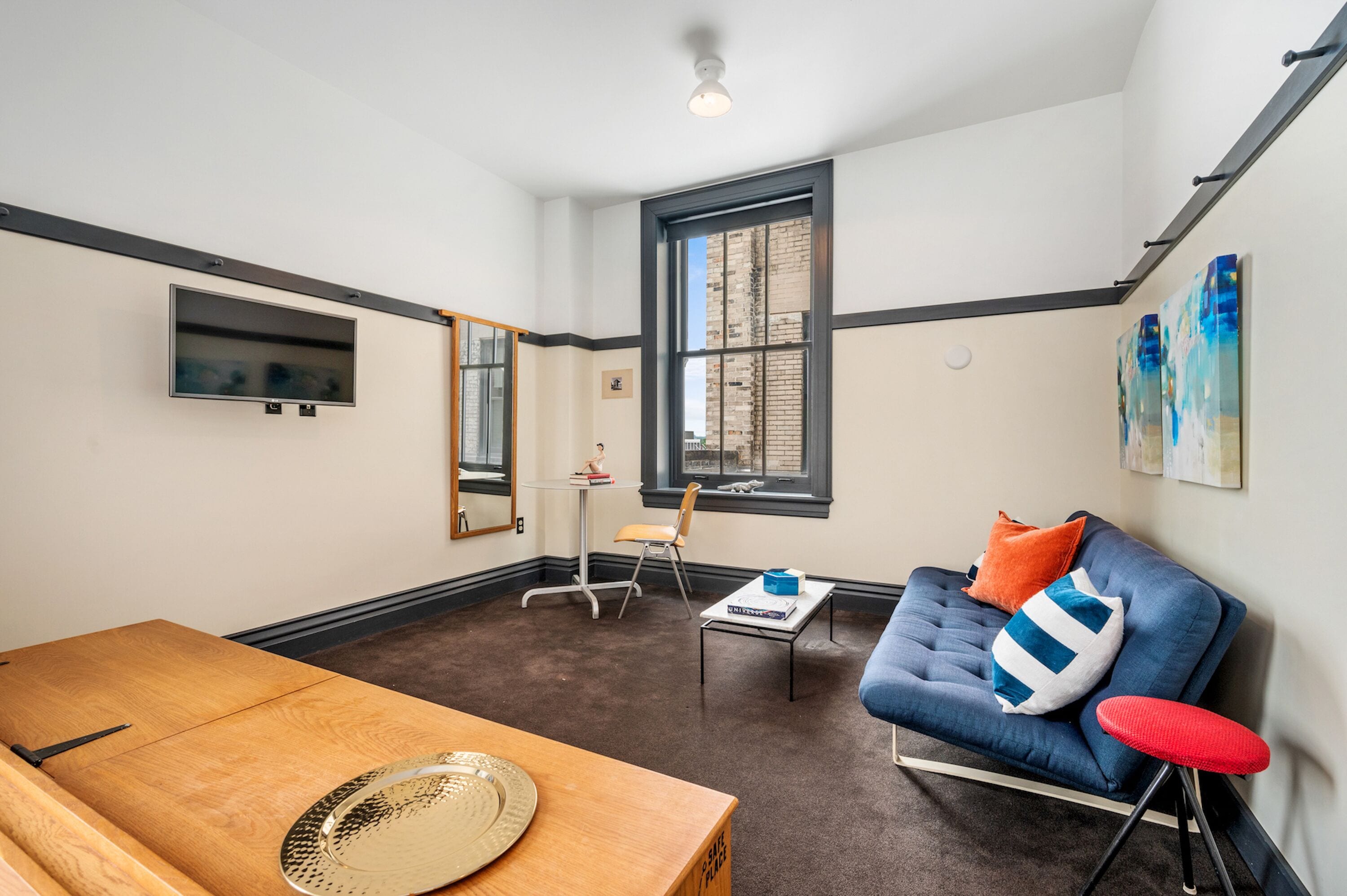 A room with a wooden table, a blue sofa with colorful cushions, a small table with books, a mirror, and a window with a view of a brick building.