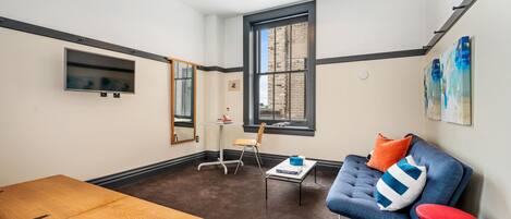 A room with a wooden table, a blue sofa with colorful cushions, a small table with books, a mirror, and a window with a view of a brick building.