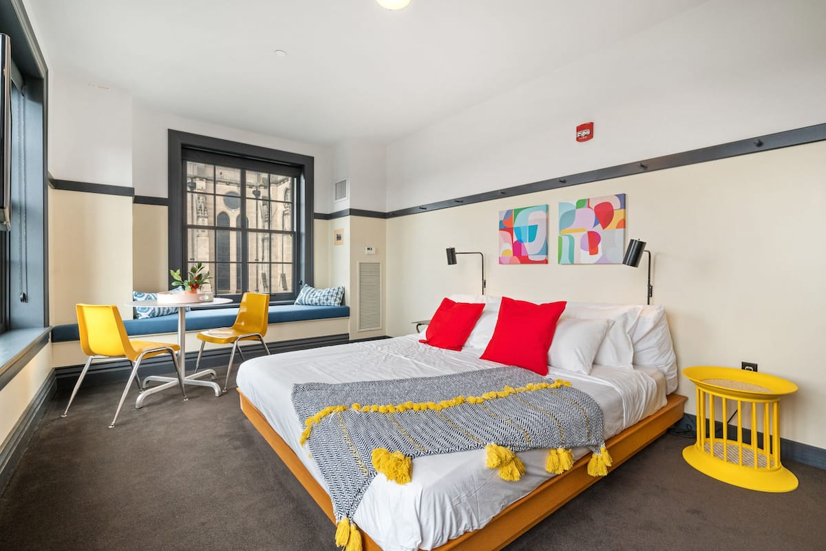 A hotel room with a bed, a desk, two chairs, a window, and artwork on the wall.