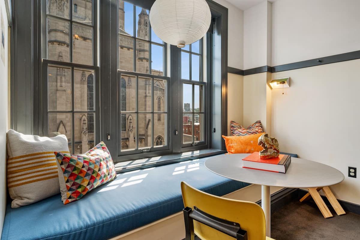 A room with a large window, a round table, and a blue cushioned bench.