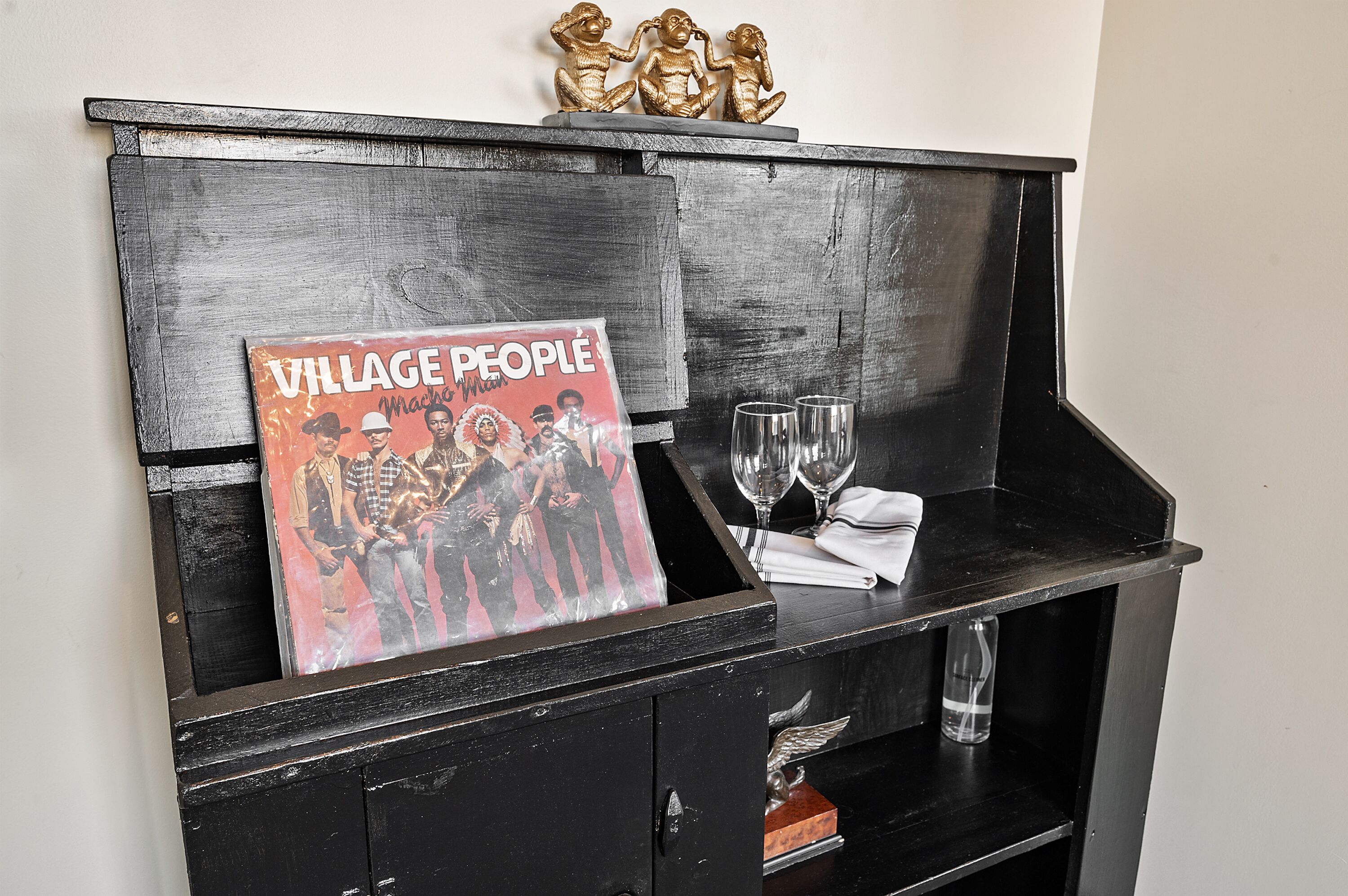 A black wooden cabinet with a vinyl record titled "Village People" and two wine glasses on top.