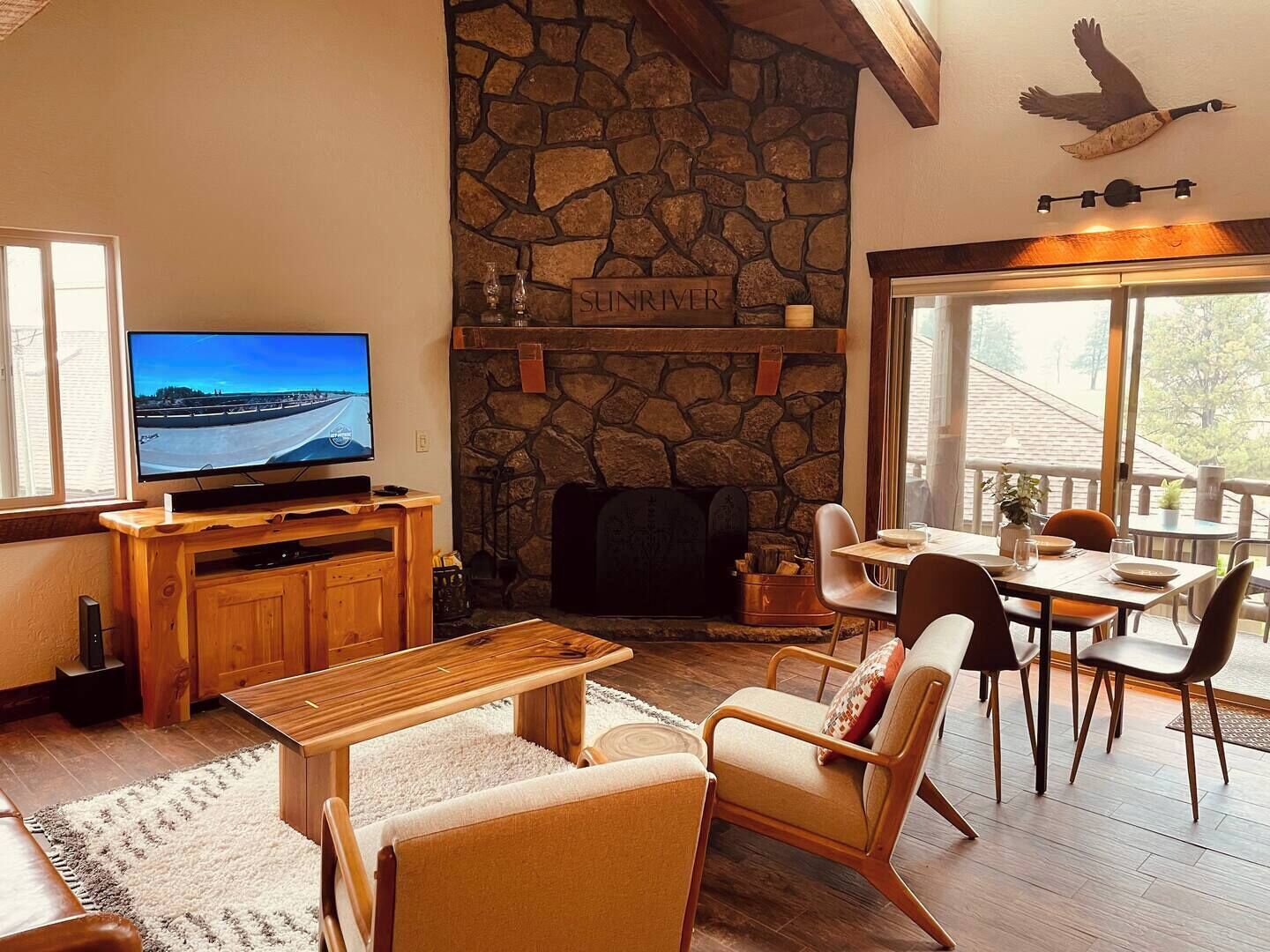 A cozy living room with a stone fireplace, a television, wooden furniture, and a view of the outdoors.