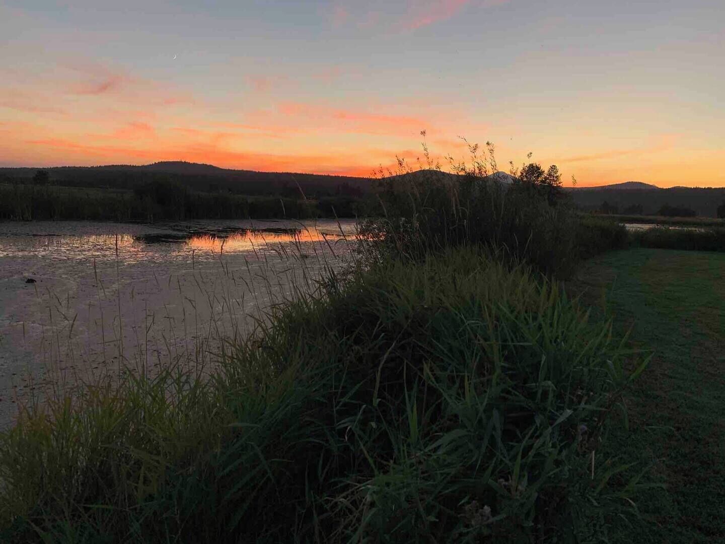 Un atardecer sobre un río con orillas cubiertas de hierba y colinas a lo lejos.