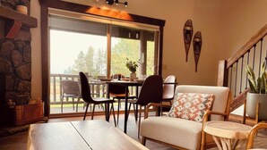 Un salon chaleureux avec une table basse en bois, un canapé et une table à manger pour quatre personnes. On peut apercevoir une cheminée en pierre et une grande fenêtre donnant sur l’extérieur.