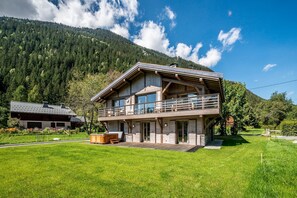 Une maison en bois à deux étages, dotée d’un balcon, entourée d’une pelouse verdoyante et bordée par une montagne boisée.