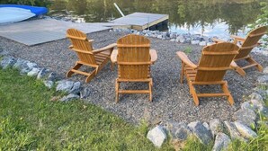 Trois chaises Adirondack en bois, disposées face à un lac paisible, avec un quai et un radeau gonflable bleu.