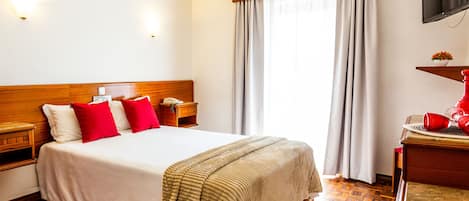 Una habitación de hotel con cabecera de madera, una cama con ropa blanca y almohadas rojas, una mesita de noche con una lámpara, una mesita auxiliar de madera, un televisor montado en la pared y una ventana con cortinas.