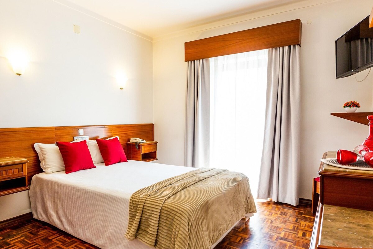 A hotel room with a wooden headboard, a bed with white and red bedding, a bedside table with a lamp, a television mounted on the wall, and a window with curtains.