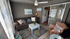 A cozy living room with a brown sofa, a white coffee table, and a wicker chair. A ceiling fan and a painting are visible on the wall. - 3's Company-"Villa Janet"- 1 Block from Gulf of Mexico, 300 feet to the Gulf (Englewood)