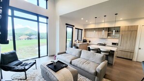A spacious living area with a grey sofa, a black chair, and a white rug. A kitchen with wooden cabinets and a dining area is visible in the background. - Perfect Hot Tub, Seconds from Skiing, Heated pool! (Park City)