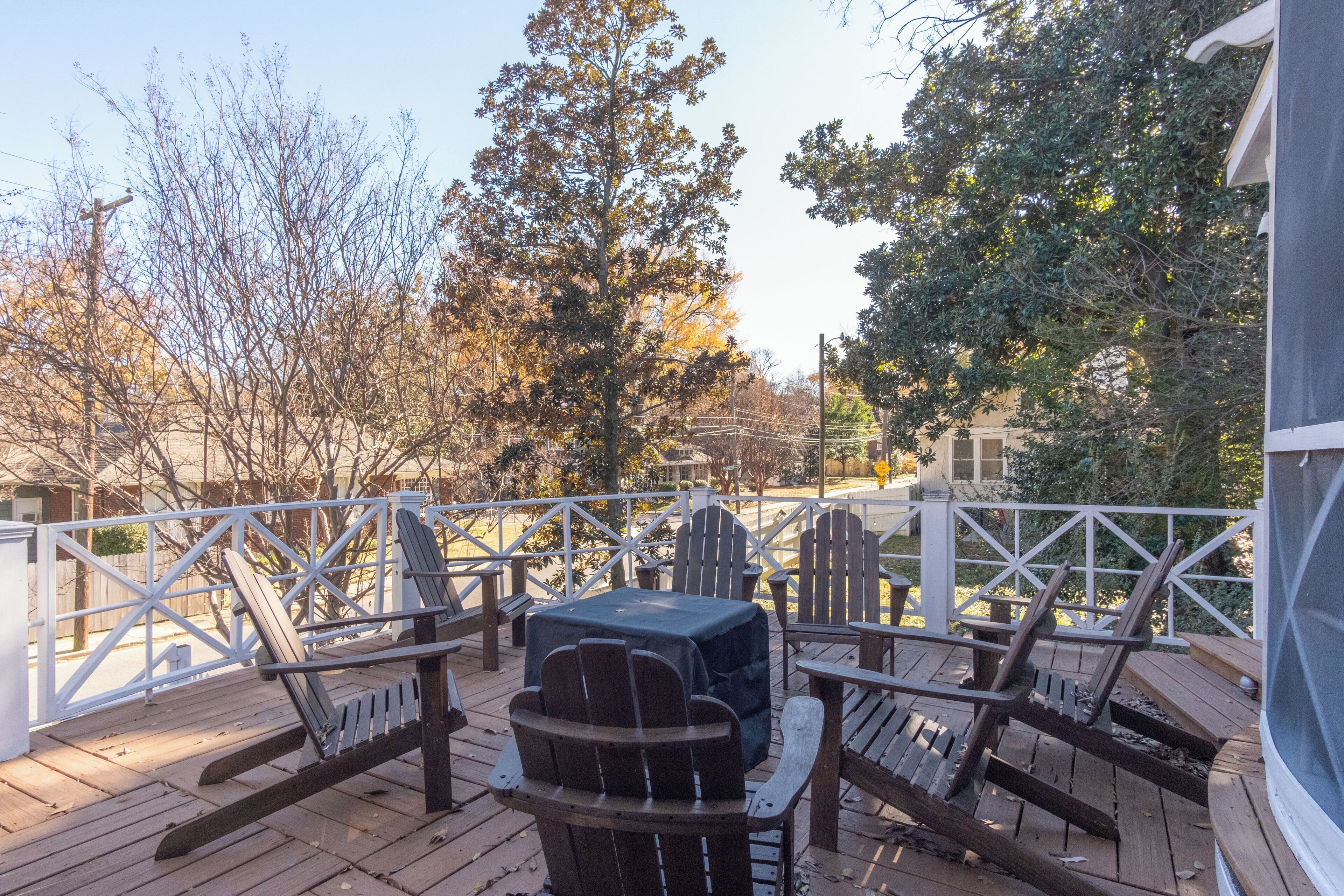 Une terrasse en bois avec des sièges d’extérieur et un barbecue, entourée d’arbres et d’une clôture blanche.