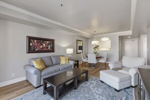 Ein modernes Wohnzimmer mit grauem Sofa, einem Couchtisch und einem weißen Sessel. Ein Essbereich mit Tisch und Stühlen ist ebenfalls vorhanden, und im Hintergrund ist eine Küche zu sehen.