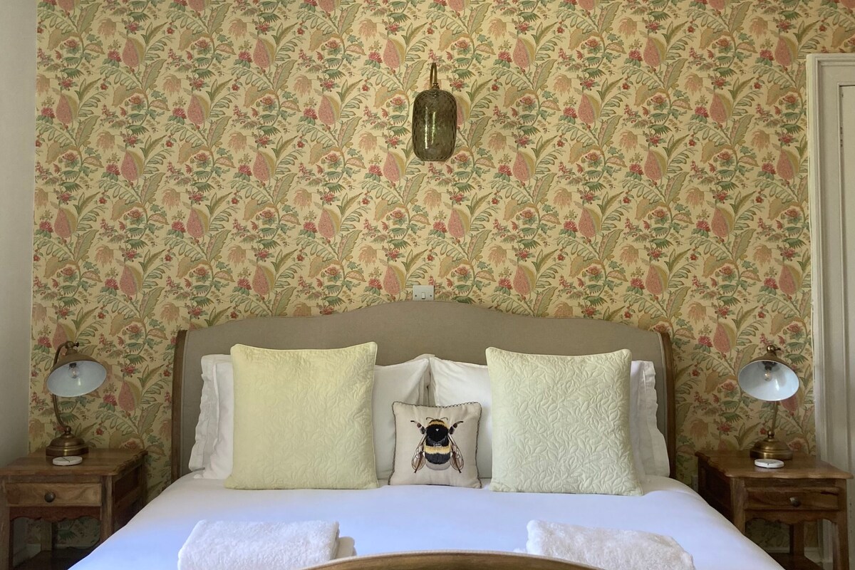 Una cama con papel tapiz floral, dos mesitas de noche con lámparas y un adorno de pared.