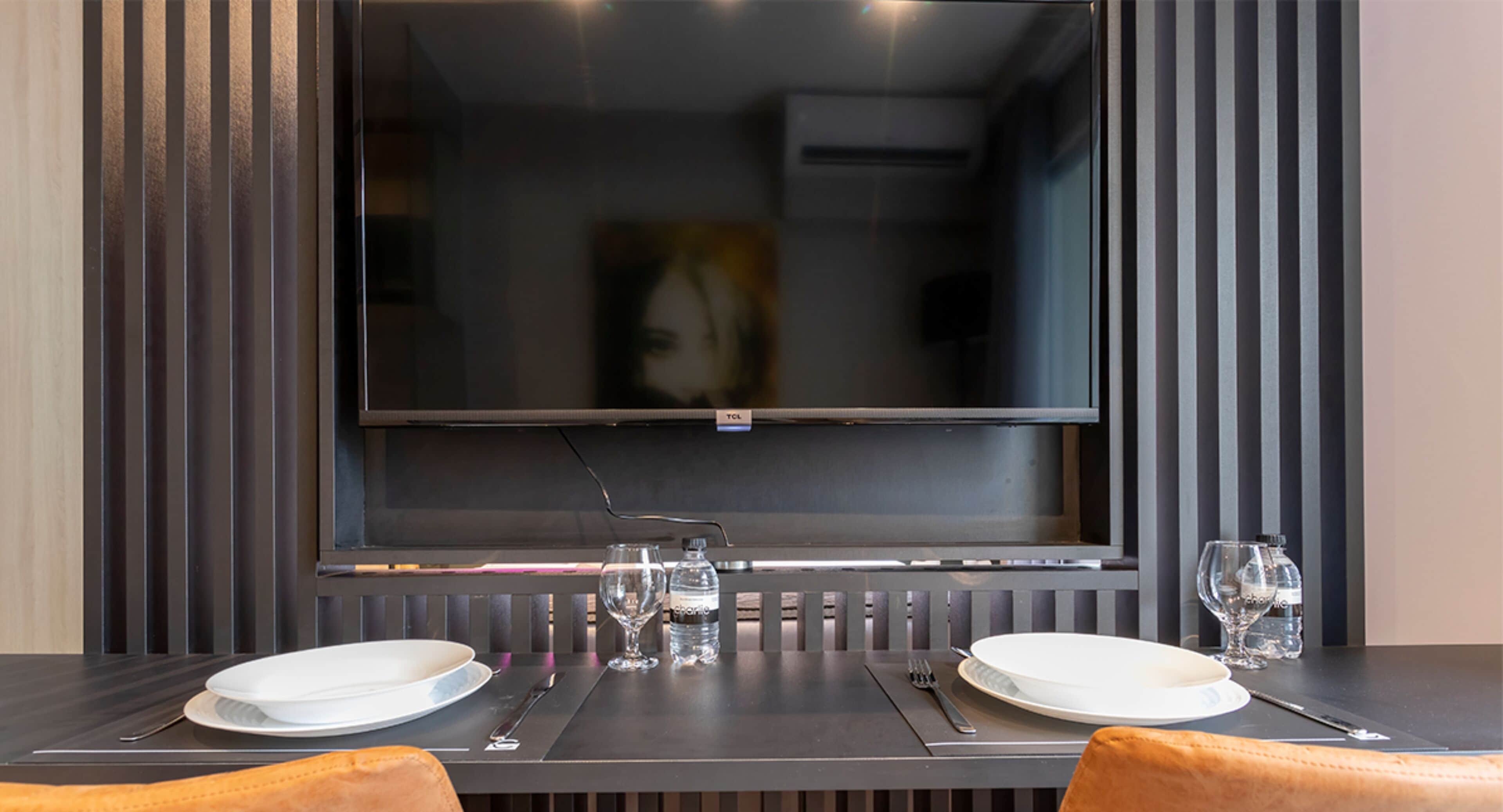 A dining area with a table set for two, a flat-screen TV mounted on the wall, and a painting above the TV.