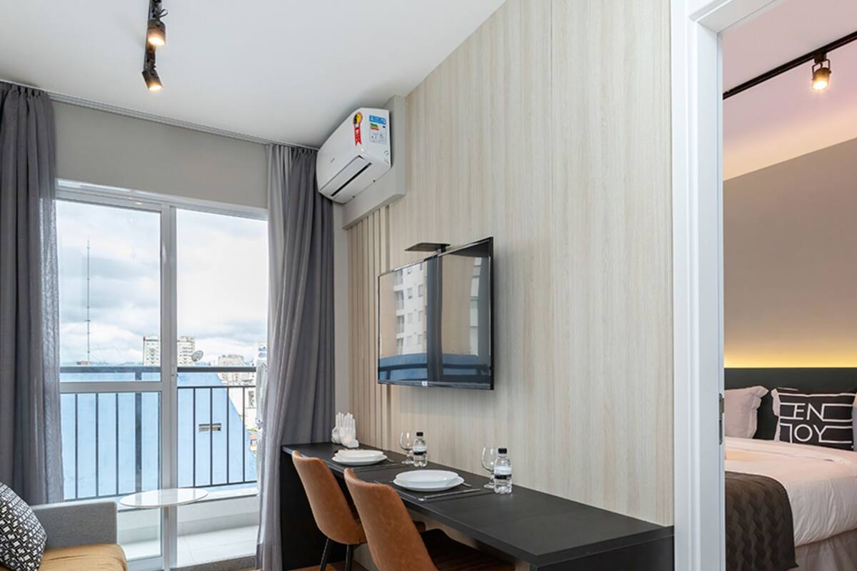 A modern hotel room with a balcony, a dining area with a table and chairs, a bed with a neon sign, and an air conditioning unit.