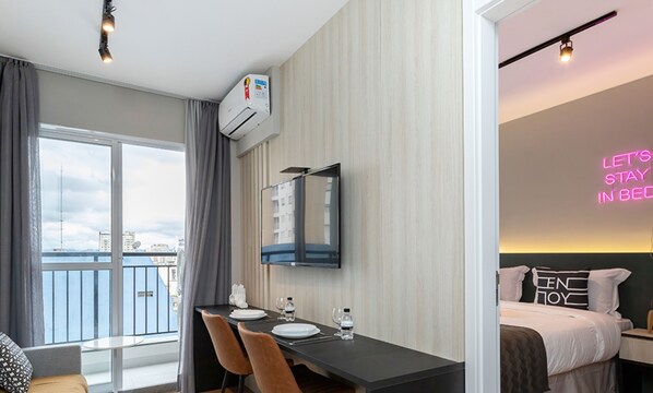 A modern hotel room with a balcony, a dining area with a table and chairs, a bed with a neon sign, and an air conditioning unit. - Smart Charlie Consolação (São Paulo)