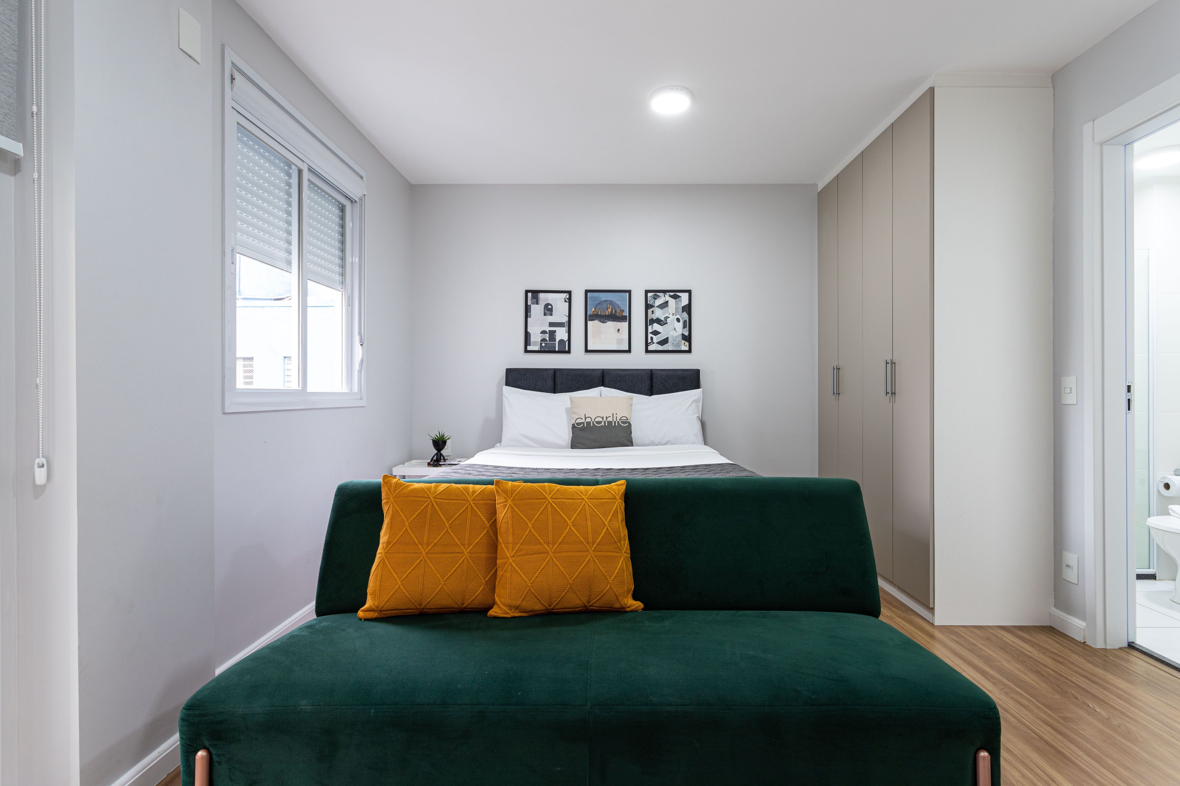 Une chambre à coucher avec un lit, un canapé vert, une fenêtre avec des stores et trois tableaux encadrés accrochés au mur.