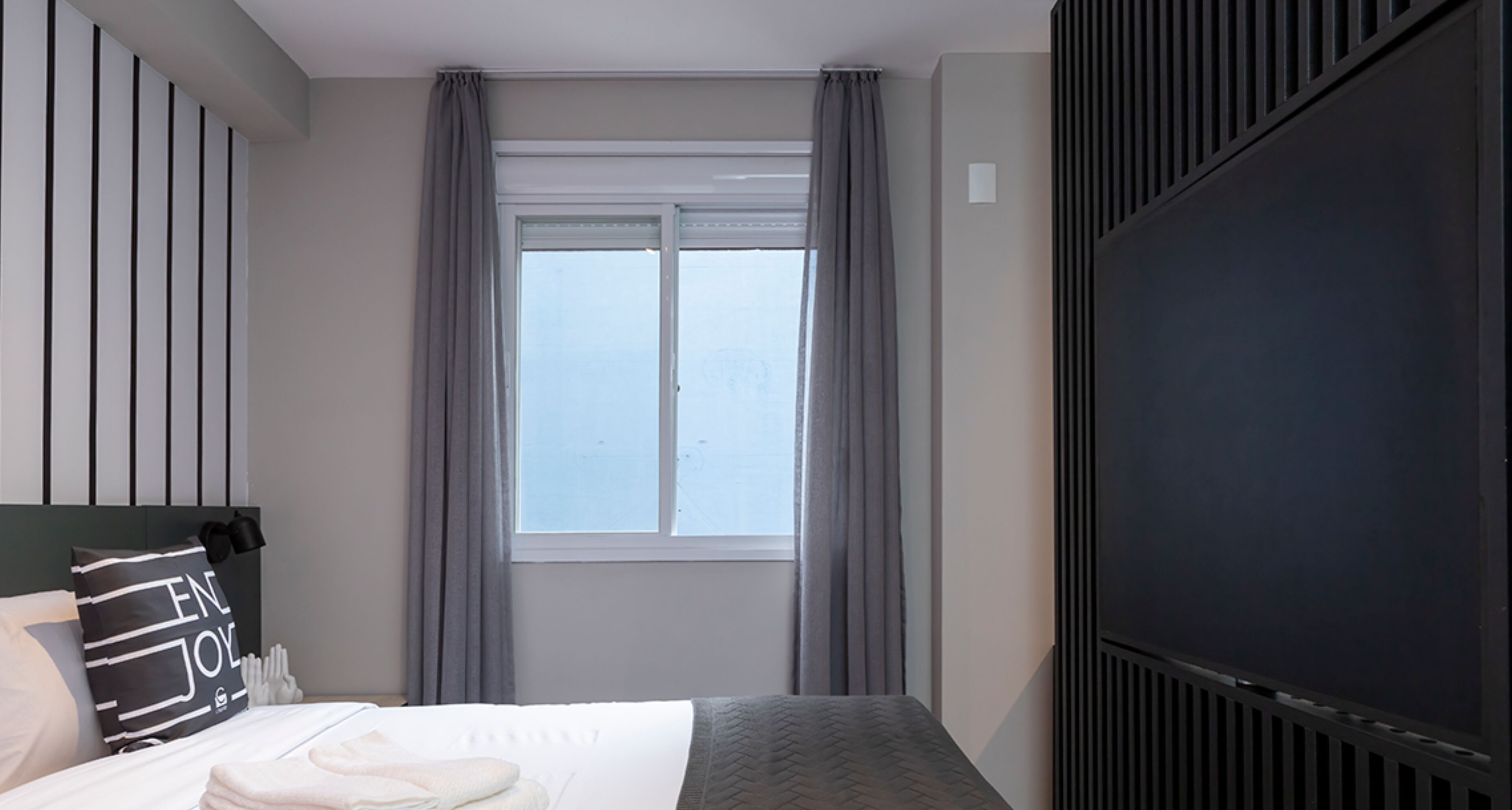 Een hotelkamer met een bed, een raam met gordijnen, een flatscreen televisie en een hoofdeinde met verticale lijnen.