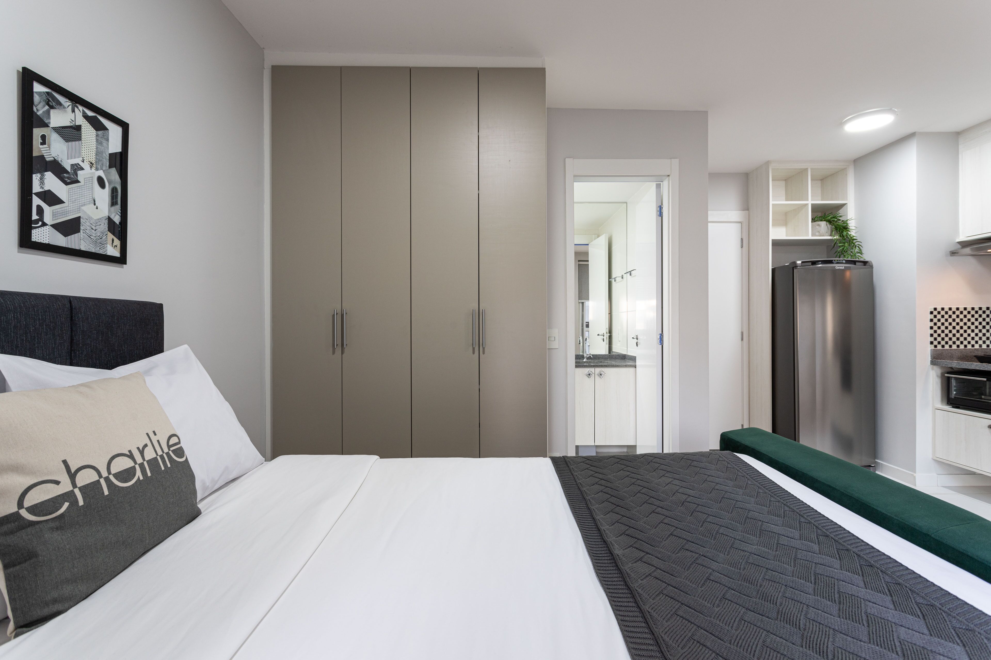 A modern hotel room with a bed, a wardrobe, a kitchenette, and a bathroom visible through an open door.