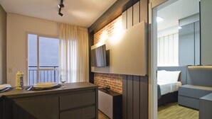 Habitación de hotel compacta con kitchenette, cama con cabecera gris, televisor montado en la pared y balcón con vistas.