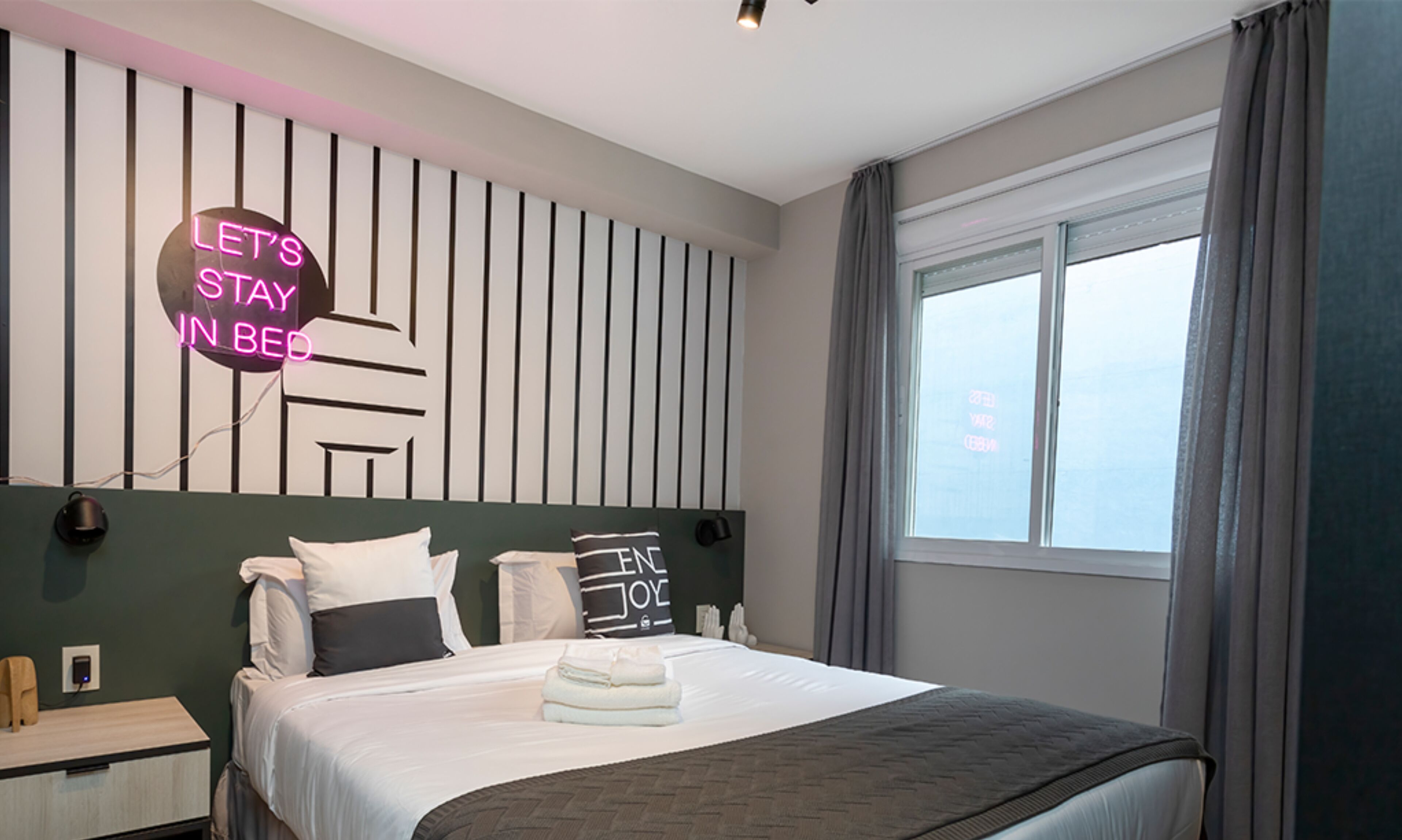 Camera d'albergo moderna con un letto, un comodino, una lampada a parete, un neon e una finestra con tende.