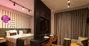 A modern hotel room with a bed, a desk, and a seating area. There is a neon sign on the wall and a large window with curtains. - Smart Charlie Consolação (São Paulo)