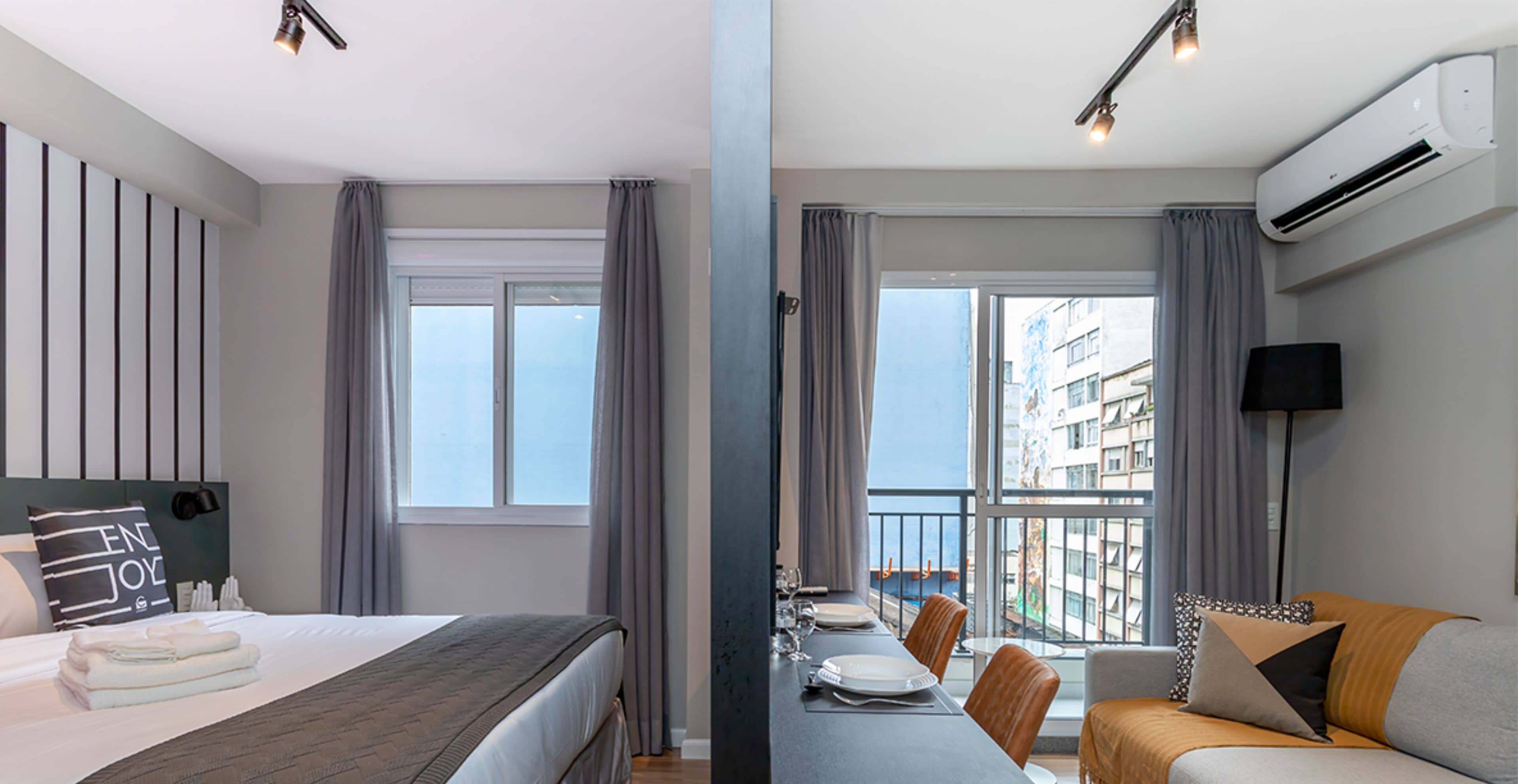 A hotel room with a bed, a dining table with chairs, a balcony with a view, and an air conditioning unit.