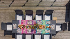 Une table à manger recouverte d’une nappe colorée, entourée de chaises en osier, dressée pour un repas avec divers plats et agrémentée d’un centre de table floral.