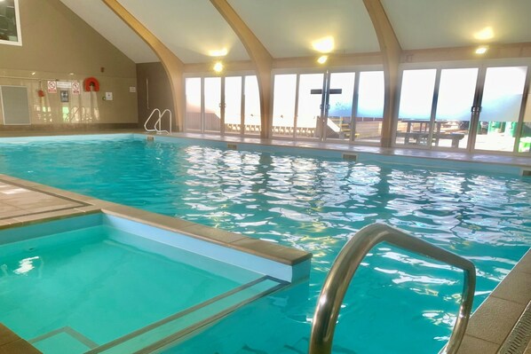 An indoor swimming pool with clear water, a diving board, and a view of the outside through large windows.