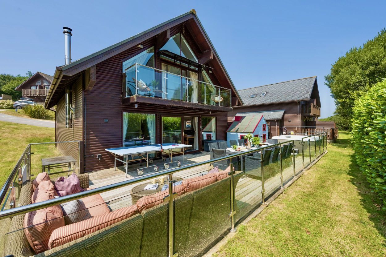 Una casa moderna de dos pisos con balcón de vidrio, mobiliario exterior y una chimenea visible.
