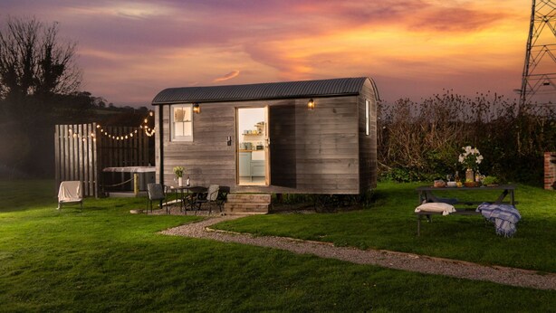 Une petite cabane en bois d’un seul étage, avec une cuisine visible à travers la porte ouverte, une terrasse avec des meubles d’extérieur et une cour gazonnée.
