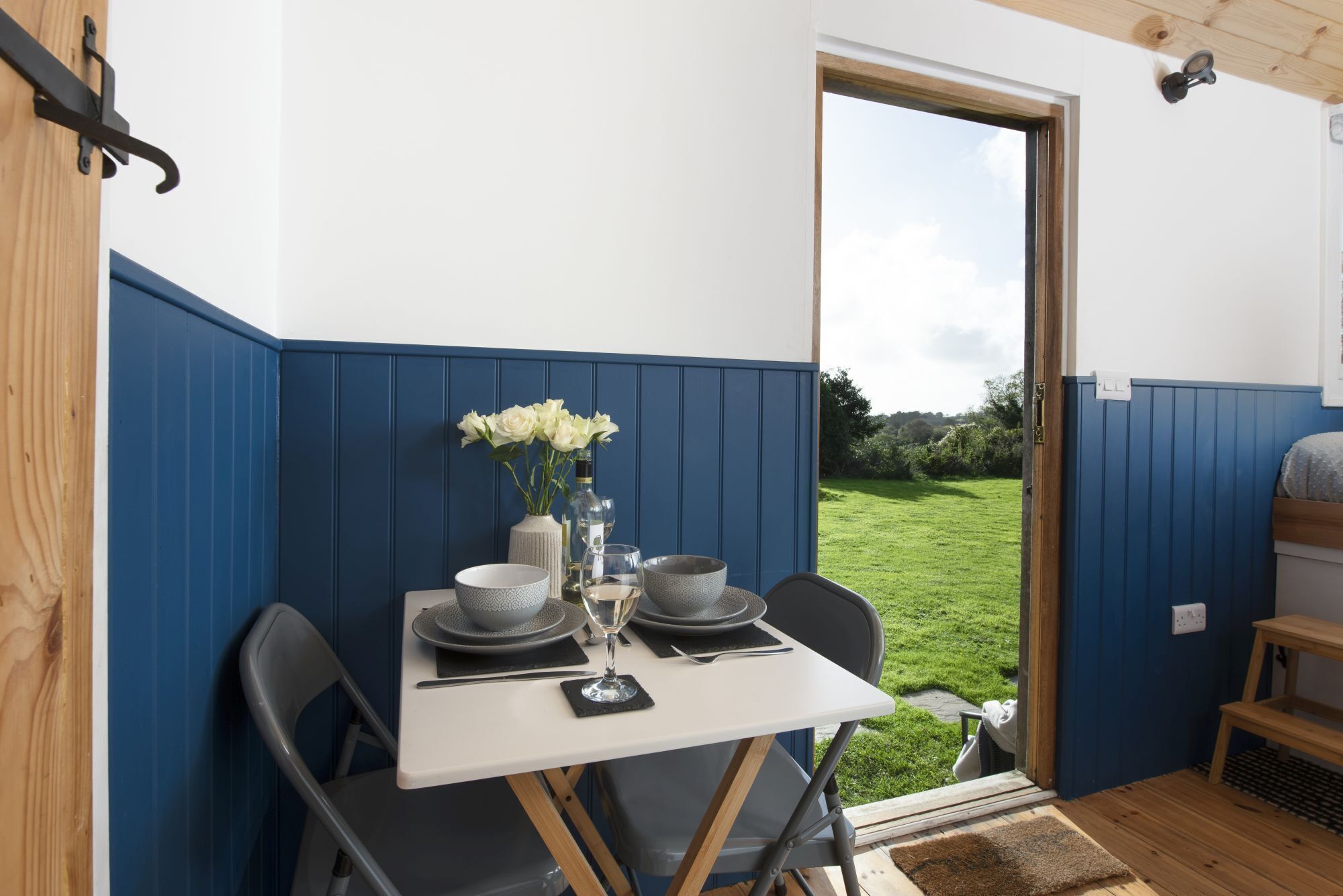 A dining area with a table set for two, a vase with flowers, and a view of a grassy landscape through an open door.