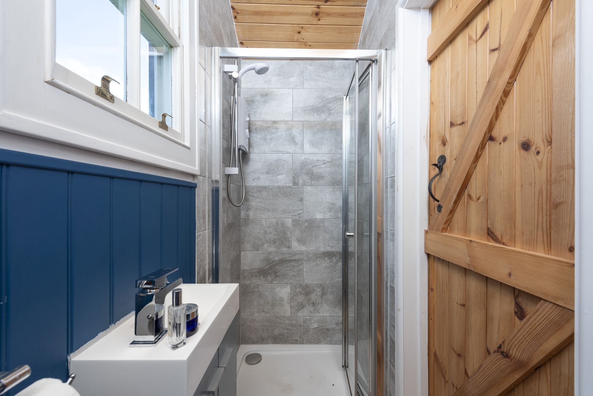 A narrow bathroom with a shower, a white sink with a mirror, and a wooden door.