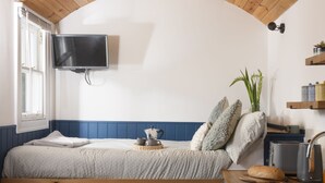 Une chambre avec une tête de lit en bois, un lit recouvert d’une couette blanche, une table de chevet avec une bouilloire, une plante et un téléviseur fixé au mur.