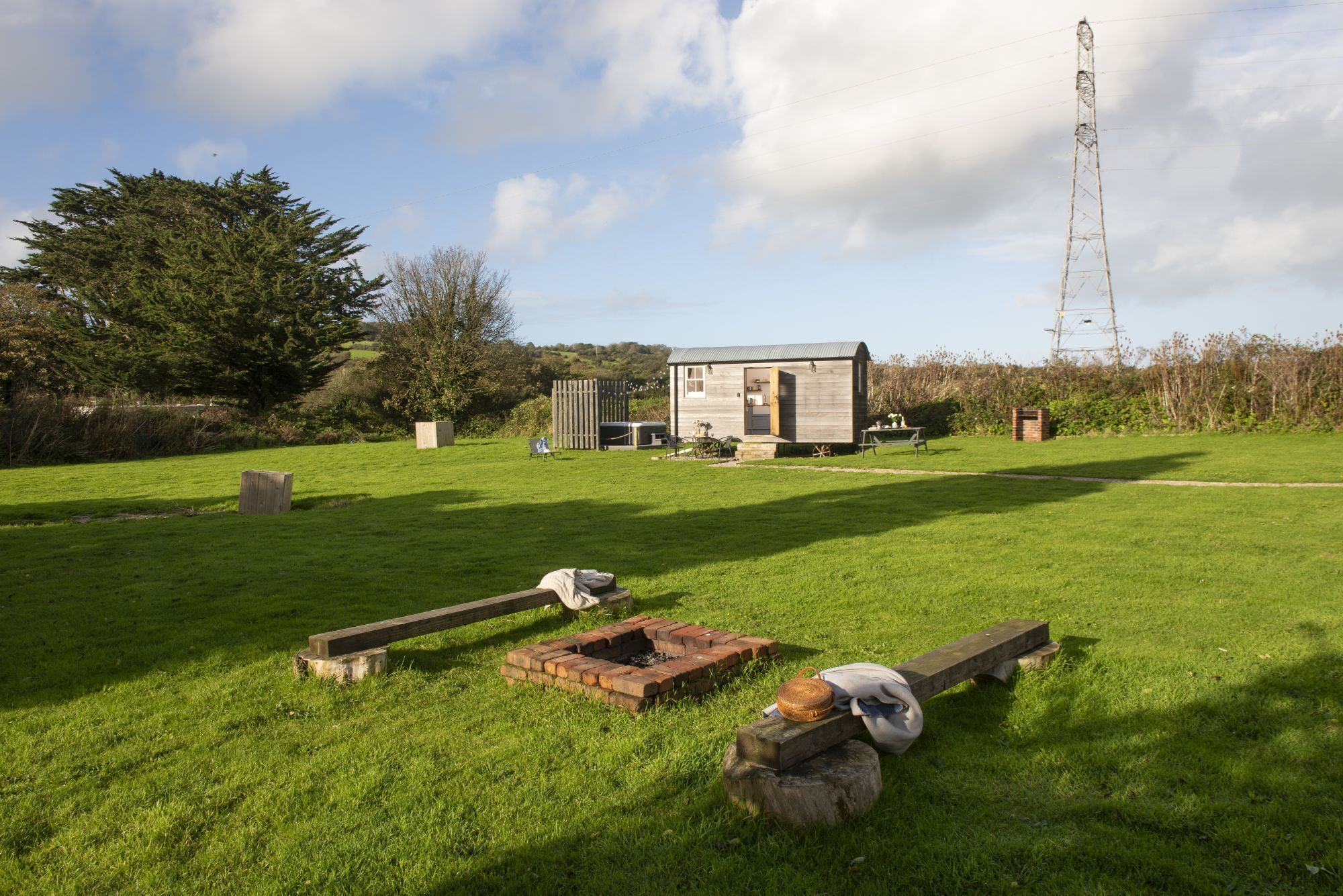 A rustic cabin with a small fire pit in a grassy field, surrounded by trees and a power line tower in the background.