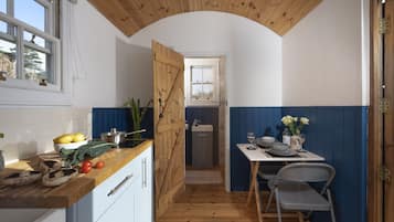 A compact kitchen with wooden ceiling, a dining area with a table and chairs, and a view through an open door to a bathroom.