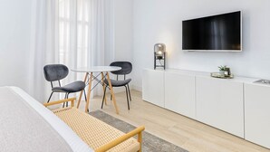 Una habitación moderna y minimalista con una cama, una pequeña mesa de comedor con sillas, un televisor montado en la pared y un armario blanco.