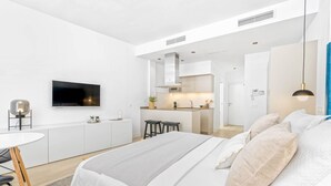 Un dormitorio moderno y minimalista con una cama grande, un escritorio pequeño y una kitchenette al fondo.