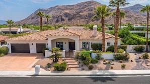 A single-story house with a tiled roof, a garage, and a well-maintained garden with palm trees and shrubs. - Luxury Estate Home in Andreas Hills All En Suite, Saltwater Pool & Spa, Fire Pit (Palm Springs)