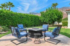An outdoor patio with a round table and four chairs, surrounded by well-maintained hedges and palm trees.