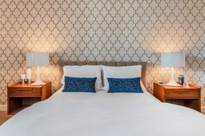 A hotel room with a patterned wallpaper, a large bed with white and blue pillows, and two bedside tables with lamps and decorative items.