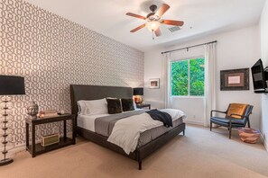 Quarto com papel de parede estampado, cama com roupa de cama branca e escura, ventilador de teto, janela com cortinas e uma cadeira.