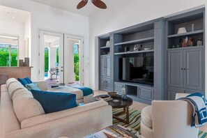 A modern living room with a sofa, armchairs, a coffee table, and a TV. There is a bookshelf and a door leading to an outdoor area.