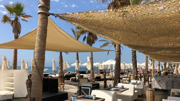 Un espace de détente aménagé sur la plage, avec des sièges blancs rembourrés, des tables et des parasols, offrant une vue sur la mer.