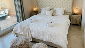 Un lit bien fait, avec du linge de lit blanc et des oreillers, deux lampes de chevet et une vue sur un espace extérieur aménagé, visible à travers une porte vitrée.