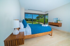 A modern bedroom with a large bed, a bedside table with a lamp, a hanging shelf with a plant, and a view of a garden through a large window. - Sophisticated Villa with Fairway Views & Private Pool, Close to the Beach (Tamarindo)