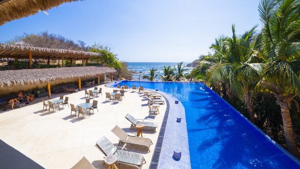 A resort pool area with lounge chairs and umbrellas, a thatched roof structure, and a view of the ocean. - Coral Blue Huatulco All Inclusive (Santa María Huatulco)