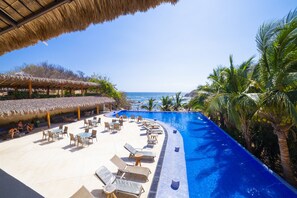 Un espace piscine aménagé dans un complexe hôtelier, avec des chaises longues et des parasols, une structure à toit de chaume et une vue sur l’océan.